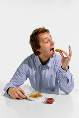 Young man about to eat crispy chicken nugget with wide open mouth and dipping sauce on table, enjoying quick meal against white background. Concept of fast food, catering, nutrition, traditional.