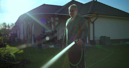 Man watering lawn with garden hose in backyard, casual moment with water spraying, residential...