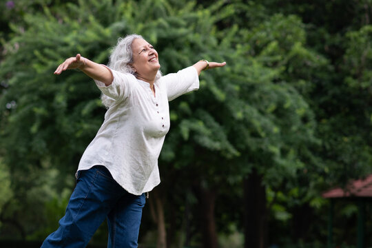 Indian happy senior woman spread her arms in environment nature like the wings of a bird. Happiness. Freedom. Elderly woman stands with one hand open in a park. Indian active old age at woman in park - Powered by Adobe