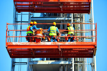 Workers in safety vests and helmets performing tasks on a construction site at an elevated platform during daylight hours