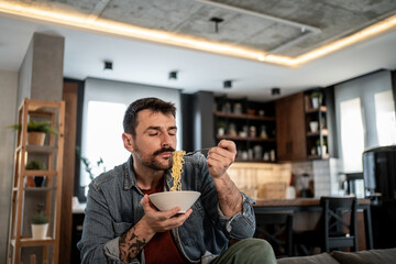 Young man savoring noodles on sofa in modern apartment