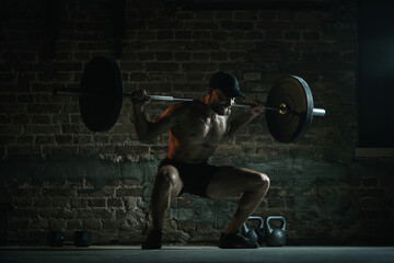 Shirtless muscular man performing deep barbell squat with loaded bar on shoulders under dramatic gym lighting with full intensity. Concept of gym culture, strength, resistance, training, healthy power