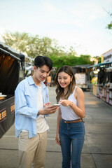 Smiling Young Asian couple looking at a smartphone while exploring a vibrant outdoor street food market, enjoying time together, and searching for places to eat