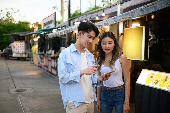 Young Asian couple deciding what to eat at a street food market, looking at a smartphone and discussing menu options from a vendor