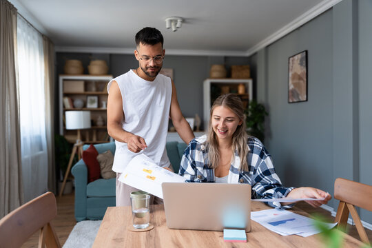Young couple working remotely at home, analyzing documents and data, using laptop in cozy modern living room, discussing financial reports and business strategy. Couple goals, simple living support.