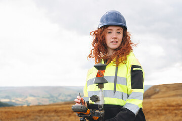 Portrait of confident female land surveyor with long hair in bright safety gear using surveying...