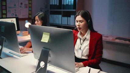 Professional asian businesswoman wearing red blazer working late at night, communicating through headset while typing on computer at dark office desk - Powered by Adobe