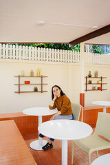 Joyful brunette curly woman in trendy pants and shirt smiles and looks out to view Pretty lady in jeans sits on chair in cafe