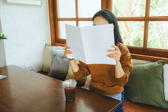 Female covering her face with blank book or magazine and sitting in chair. - Powered by Adobe
