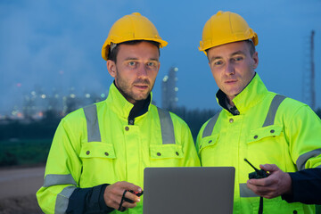 Two engineers in safety gear collaborate at dusk near construction site with city lights in background