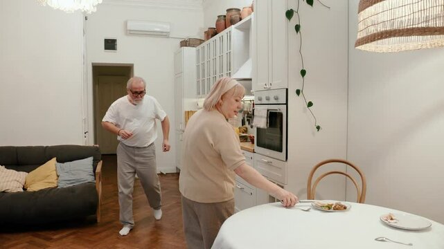 Grandma takes a bottle and cutlery from the table, then leaves and grandfather approaches, taking plates from the table