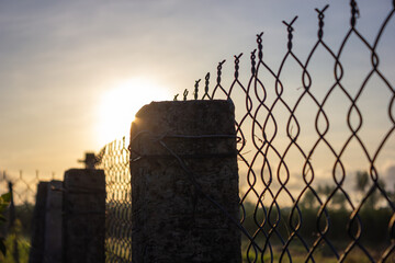 Perspective view of a chain link fence and concrete posts silhouetted against a bright sunset or...
