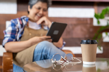 Takeaway coffee cup and eyeglasses placed on glass table in foreground with smiling Asian senior woman in apron using tablet in background, showing relaxed lifestyle and digital cafe environment.