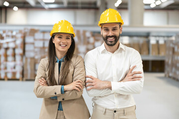 Man and woman with yellow safety helmets posing in warehouse with arms crossed.