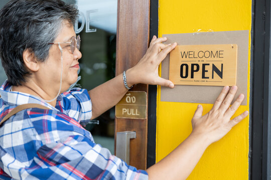 Asian senior woman wearing plaid shirt and apron putting open welcome sign on yellow wall in front of cafe door. Close-up moment showing pride, confidence, and readiness to run local business. - Powered by Adobe