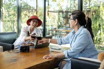Two Asian senior women sitting in cafe toasting red wine glasses with joyful smiles, laptop and camera on wooden table, enjoying relaxed conversation and celebrating friendship in cozy modern setting.