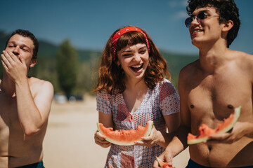 Group of friends laughing and enjoying slices of watermelon at the beach on a sunny day. A depiction of friendship, summer fun, and happiness outdoors near the lake.