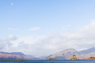 Castle Stalker bei Tageslicht vom See aus gesehen, sonnig beleuchtet mit sichtbarem Mond am Himmel,...