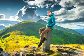 Man resting on a mountain peak in soft sunlight, wearing a green jacket and brown hiking pants,...