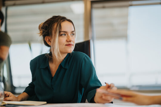 A confident businesswoman in a teal blouse sits attentively during a meeting. The setting conveys a professional and collaborative atmosphere, emphasizing focus and engagement.