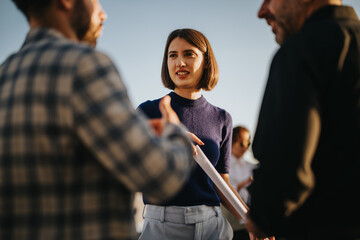 Plakat A diverse group of business colleagues engage in a conversation on a high tower balcony during sunset. The scene captures a collaborative and professional atmosphere with a multicultural team.