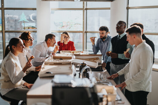 A diverse group of business employees gathers around a table enjoying pizza during a casual lunch break, fostering teamwork and collaboration in a modern office environment.