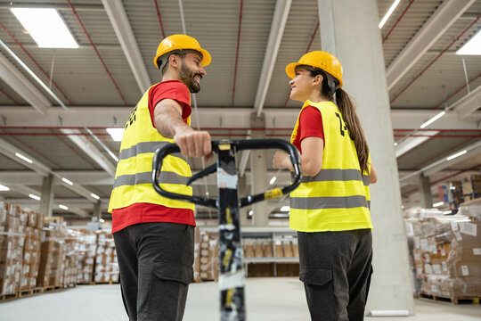 Man and woman in safety vests and helmets talking beside manual pallet jack inside warehouse. - Powered by Adobe