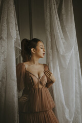 Elegant woman in a brown dress posing near white curtains in a graceful studio setting