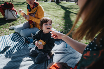 A young child enjoys a day outdoors while having a picnic on a sunny day with family, surrounded by...