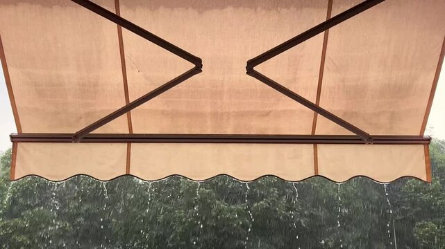 Summer rain falling on green trees, view from cozy city terrace with classic Italian awning. Calm urban atmosphere with soft raindrops and lush foliage