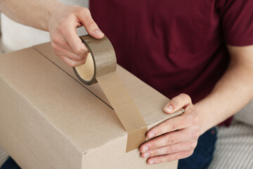 Man sealing cardboard box with adhesive tape indoors, closeup