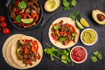 Fajitas, beef meat slices with colored pepper and onions, traditional mexican food, served with tortillas, salsa and sour cream on black background, top view. High quality photo