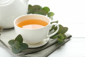 Aromatic tea in cup and eucalyptus branches on white wooden table, closeup