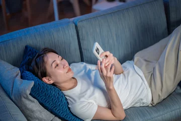Zelfklevend Fotobehang ソファに寝転んでスマホを使う女性 © naka