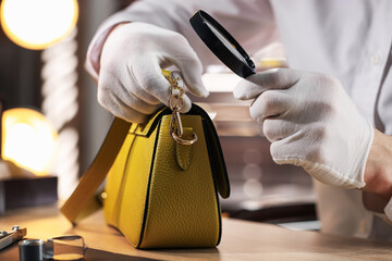 Appraiser with magnifying glass evaluating leather bag at table indoors, closeup