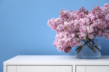 Beautiful lilac flowers in glass vase on cabinet near light blue wall, space for text