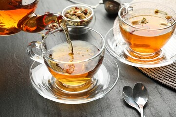 Pouring herbal tea from teapot into cup at black table, closeup