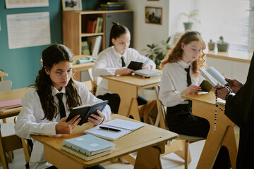 Group of high school students engaging in classroom activities while reading and studying together at desks. Teacher holding materials, supervising the class in educational setting