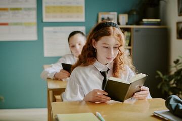 Portrait of young Caucasian schoolgirl focused on reading book while sitting at desk in classroom, with classroom charts visible in background