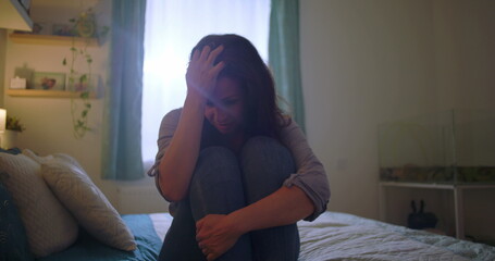 Woman sitting on bed, knees pulled close, tears in her eyes, overwhelmed with sadness, themes of...