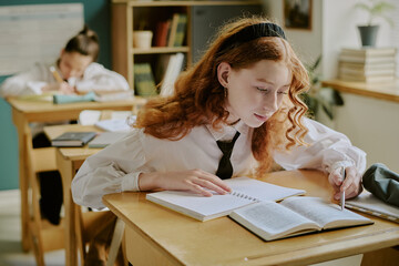 Student with red hair studying in classroom surrounded by open books, focusing on preparations for...
