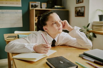 Young student sitting at desk leaning head on hand, appearing to be deep in thought while holding...