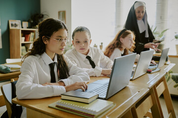 Group of students wearing uniforms sitting at desks and working on laptops with teacher dressed in religious attire supervising in warm lit classroom