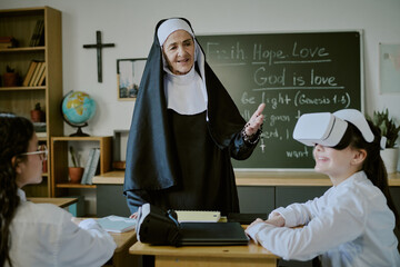 Elderly teacher in habit interacting with students using virtual reality headset in classroom setting with bookshelves and globe. Friendly education environment for immersive learning experience