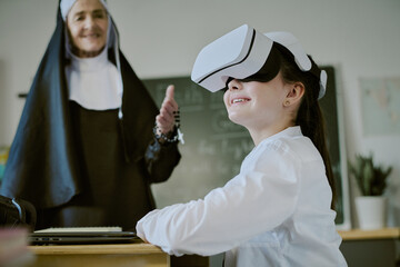 Young student wearing virtual reality headset in classroom being observed by smiling nun standing beside whiteboard and books on table