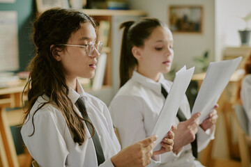 Two girls wearing school uniforms reading documents in classroom setting with shelves and books in the background and their focused expressions indicating concentration