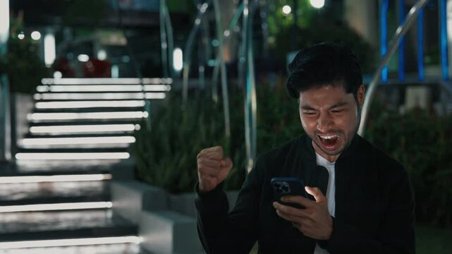 Ecstatic male celebrating triumph and screaming yes while winning online game in electronic device. Fashionable man playing gambling and making fortunate bet while standing by illuminated stairs.