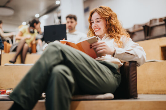A happy woman sitting comfortably while reading a book in a contemporary office space, with co-workers collaborating in the background, creating a relaxed and focused atmosphere.