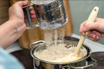 Adding sifted flour from a measuring cup into dough on a double boiler, showcasing a traditional baking technique.