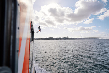 A boat navigating through a calm sea with a cityscape and bridge in the background under a cloudy sky.